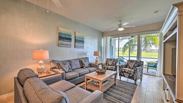 A living room with a sofa, armchairs, a coffee table, and a TV. There are ceiling fans, a lamp, and artwork on the wall.