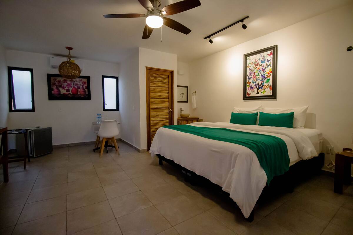 A bedroom with a bed, a ceiling fan, a desk, a chair, a small table, and a window.