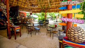 An outdoor café with tables and chairs, a thatched roof, and colorful signs pointing to destinations like Chetumal and California.