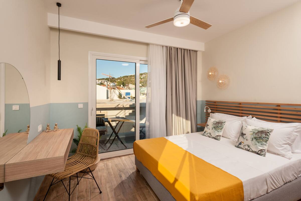 A hotel room with a bed, a desk, a chair, a ceiling fan, and a view of the outside.