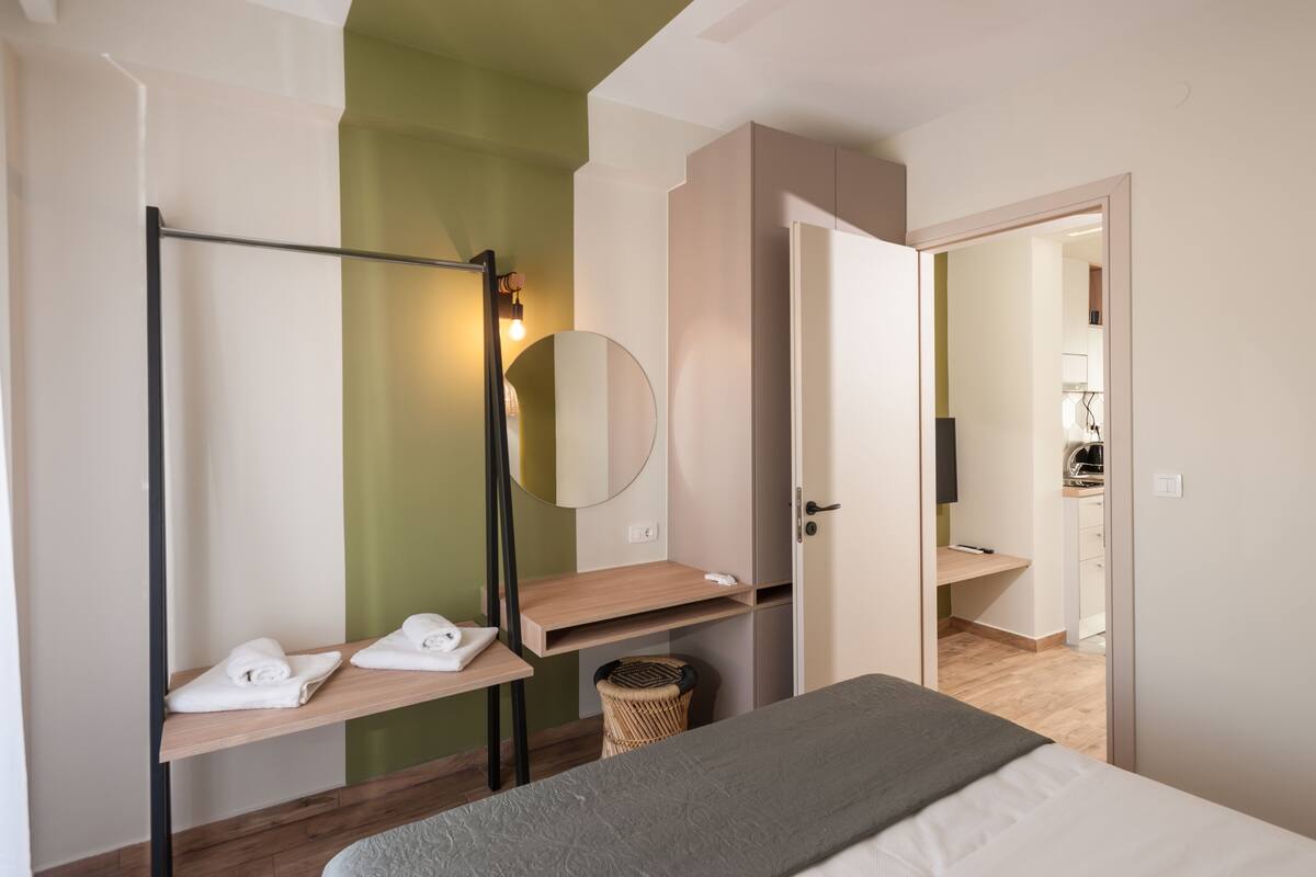A modern bedroom with a bed, a wooden shelf with towels, a mirror, and a door leading to a bathroom.