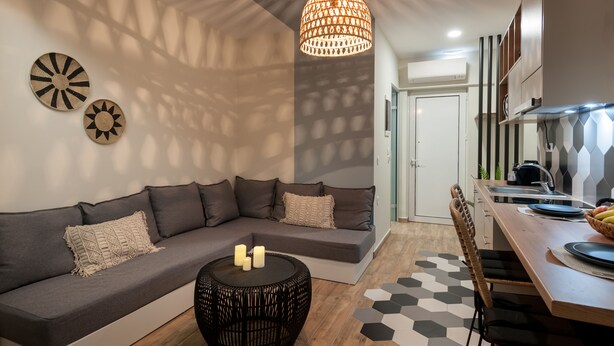 A modern living room with a grey sofa, a round coffee table with candles, and a dining area with a wooden table and chairs. - Salty Colours Apartments (Apokoronas)