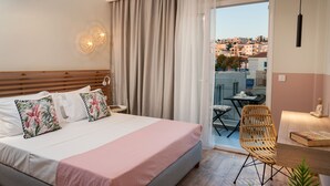 A hotel room with a bed, a chair, a table, and a view of the city through a balcony. - Salty Colours Apartments (Apokoronas)