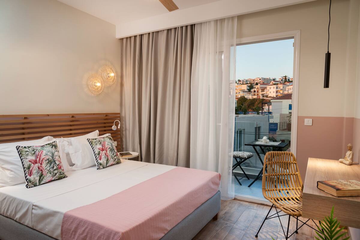 A hotel room with a bed, a chair, a table, and a view of the city through a balcony.