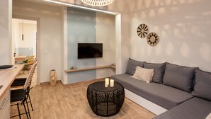 A modern living room with a sofa, a coffee table, a TV mounted on the wall, and a dining area with chairs and a table. - Salty Colours Apartments (Apokoronas)