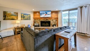 A modern living room with a stone fireplace, a large flat-screen TV, and a cozy seating area. - Lakeside Condo | Balcony, Pool + Summer Adventures (Keystone)