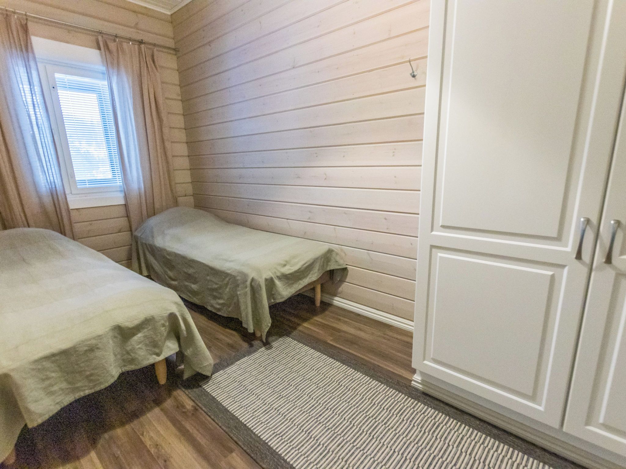 Une chambre avec deux lits simples, une fenêtre avec des rideaux, une armoire et un tapis à rayures.
