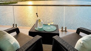 Een balkon met rieten meubilair, een tafel met fles en glazen, en uitzicht op een wateroppervlak.