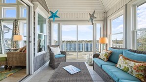 A sunlit room with wicker furniture, a sofa, and a table, featuring large windows and a view of the sea. - Compass Tres: Guiding You To Vacation Perfection! (North Topsail Beach)
