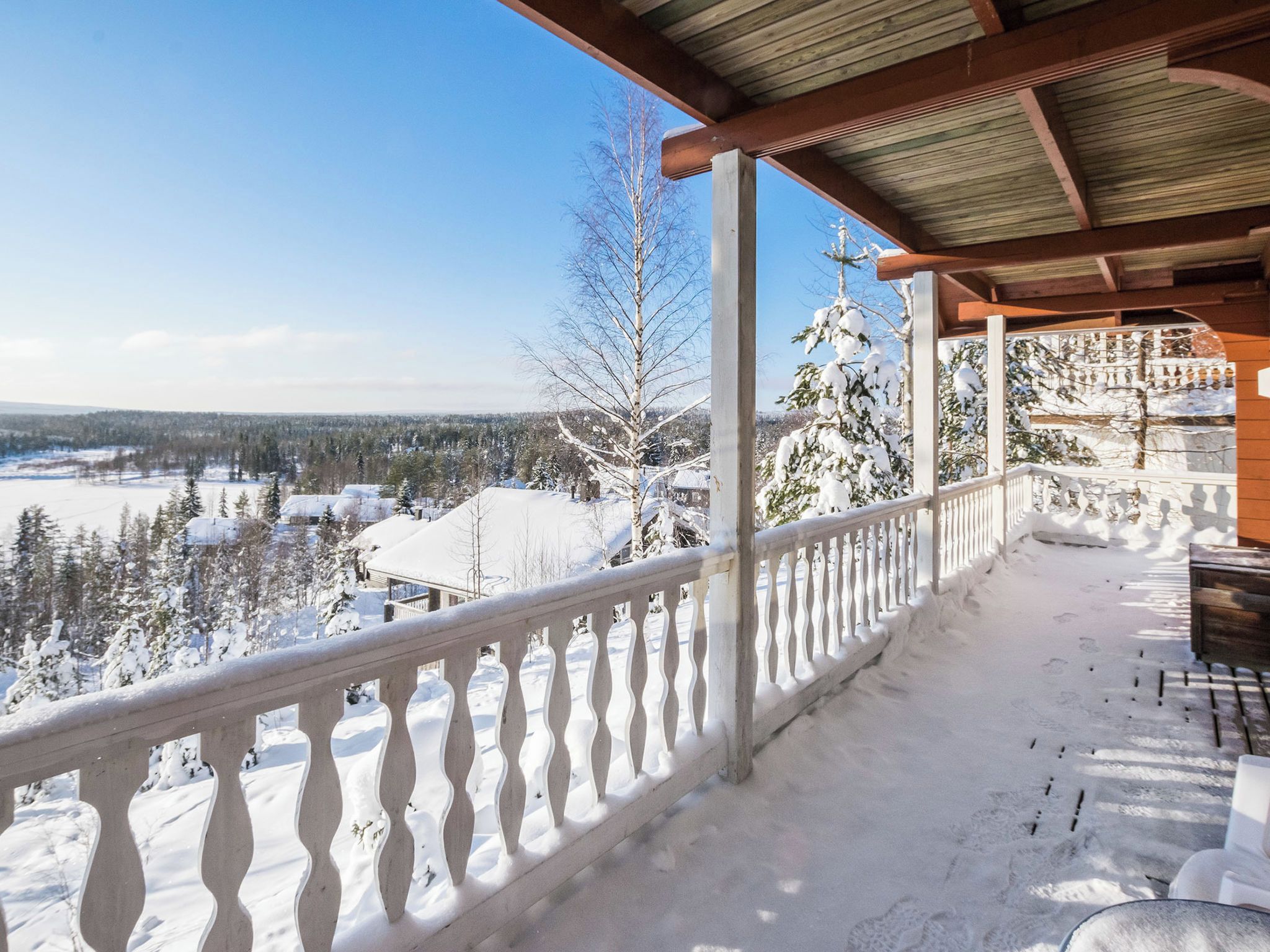 A snow-covered balcony with a wooden railing overlooking a snowy landscape and trees.