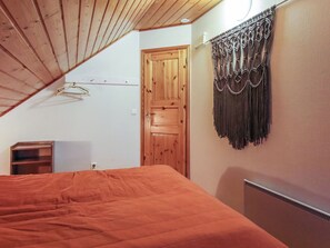 A bedroom with a sloped ceiling, a wooden door, a macrame wall hanging, and a bed with an orange bedspread.