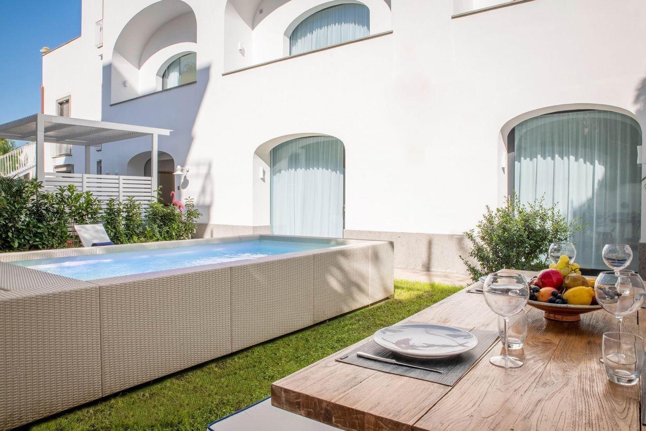 A modern outdoor pool area with a wooden deck, a dining table set with plates and glasses, and a fruit bowl.