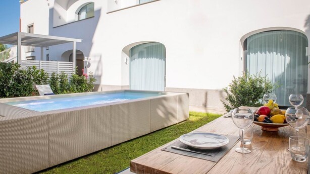 A modern outdoor pool area with a wooden deck, a dining table set with plates and glasses, and a fruit bowl. - Luxury Apartment with Garden and Pool (Sant'Agnello)