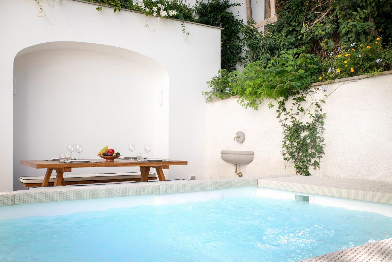 Zona de piscina amb taula de fusta preparada per dinar, paret blanca amb vegetació i un petit rentapeus.