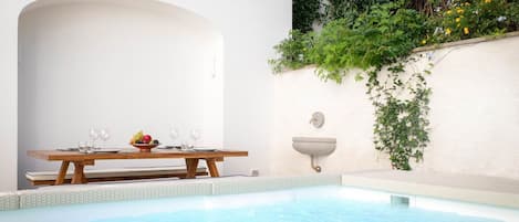A pool area with a wooden table set for dining, a white wall with greenery, and a small sink.