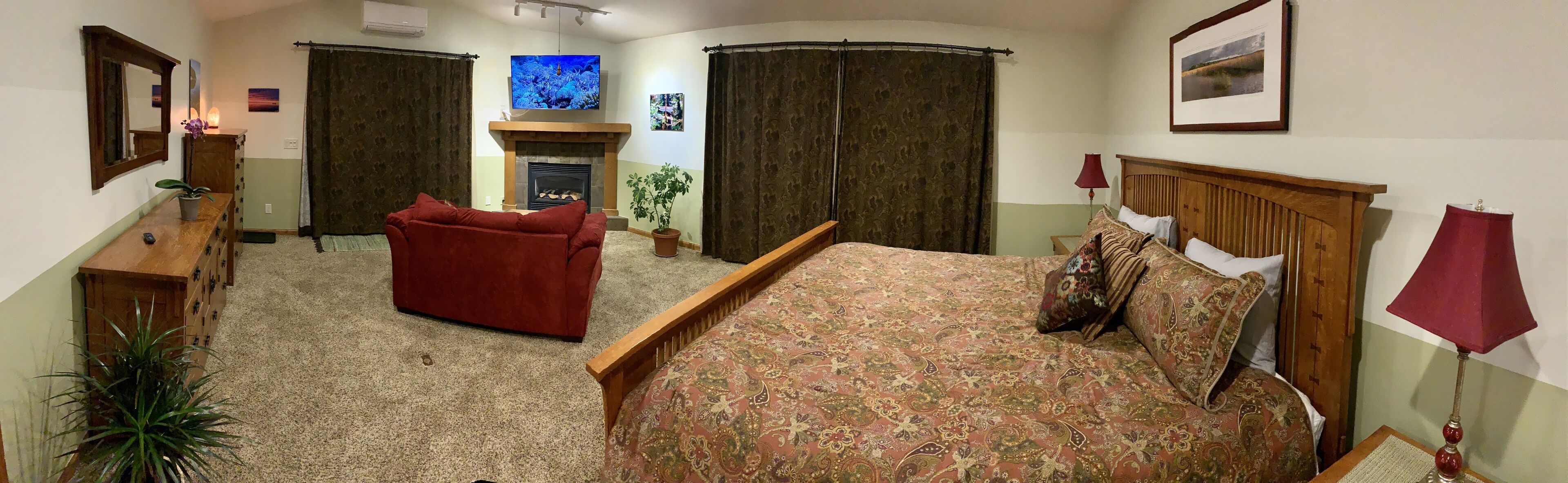 A hotel room with a bed, a red armchair, a wooden dresser, a mirror, a fireplace, and a television.