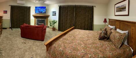 A hotel room with a bed, a red armchair, a wooden dresser, a mirror, a fireplace, and a television.