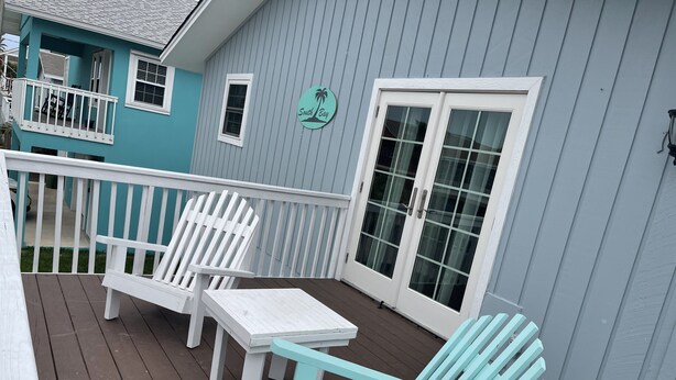 Een overdekt terras met witte Adirondack-stoelen en een kleine tafel, een blauw huis met een bord waarop 