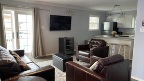 Een woonkamer met een bruine leren bank, een flatscreen televisie en een keuken met witte kasten en een barkruk.