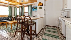 A cozy room with a wooden ceiling, a dining area with a table and chairs, a kitchen with white cabinets, and a patterned floor. - Alppikylä 13b by Interhome (Hyrynsalmi)