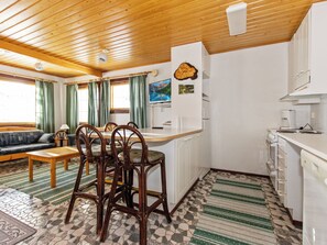 Un salon chaleureux avec un plafond en bois, un coin repas avec des chaises en osier, une cuisine avec des meubles blancs et une grande fenêtre ornée de rideaux verts.