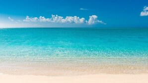 Une plage à l’eau turquoise et limpide, bordée d’un rivage sablonneux, sous un ciel bleu parsemé de nuages.