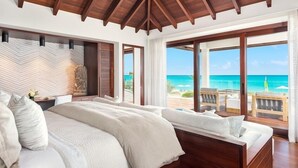 Une chambre dotée d’un grand lit, de poutres apparentes en bois au plafond, d’une vue sur la plage et d’un balcon aménagé avec du mobilier d’extérieur.