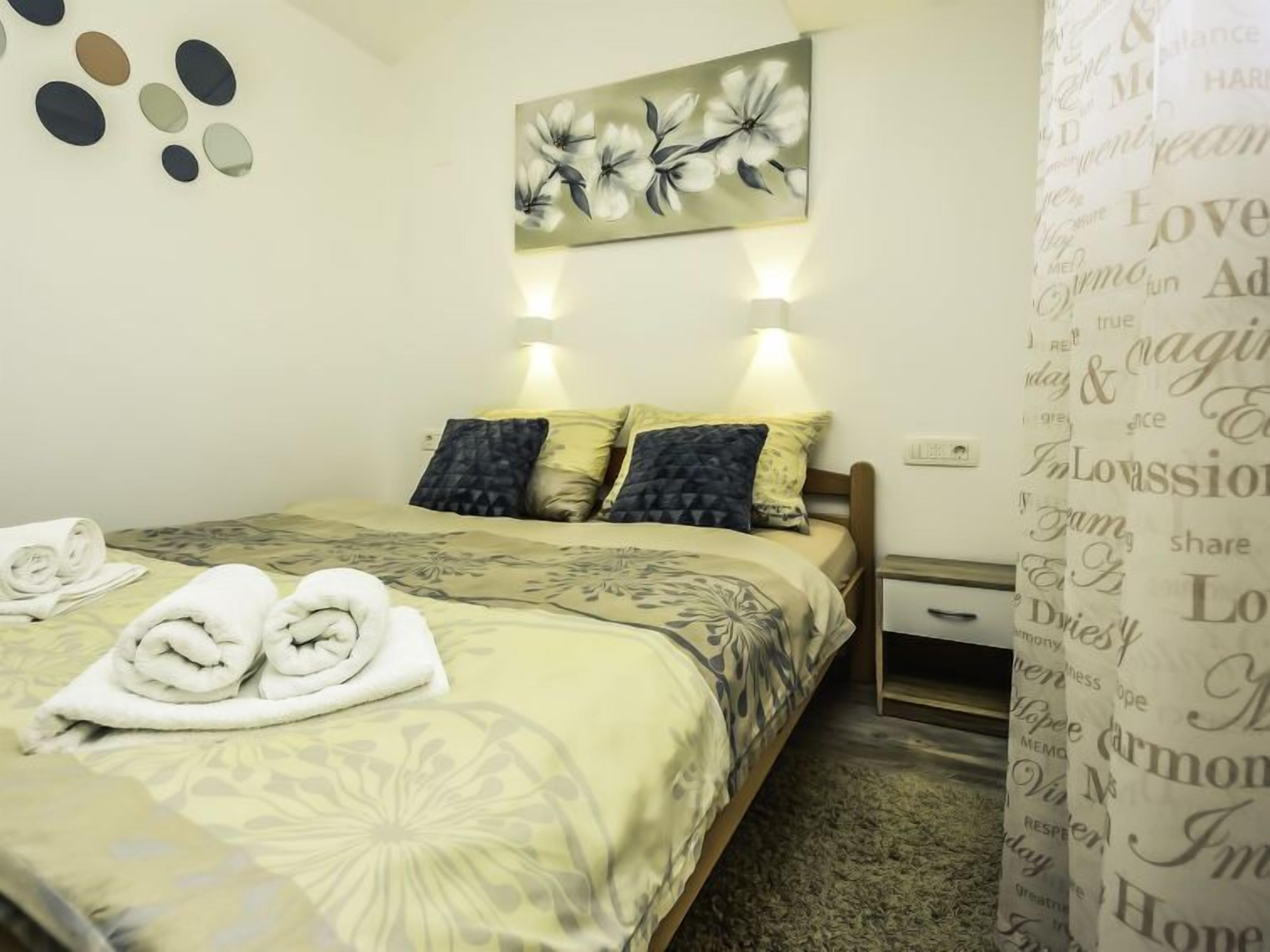 Een net opgemaakt bed met een bloemenbed overtrek en twee kussens, een aan de muur hangend bloemen schilderij, en een gordijn met tekst.