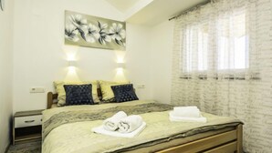 A bedroom with a bed, two bedside tables, a wall-mounted light, a window with sheer curtains, and a floral painting above the bed.