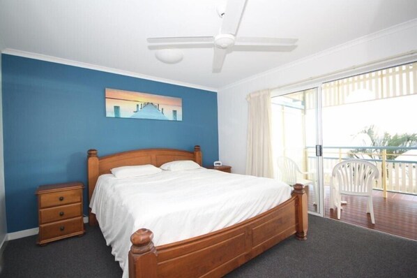 Un dormitorio con cama de madera, una mesita de noche, ventilador de techo y vista al balcón.