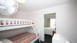 Une chambre avec un lit superposé, un ventilateur blanc, une petite fenêtre et une porte donnant sur une autre pièce.