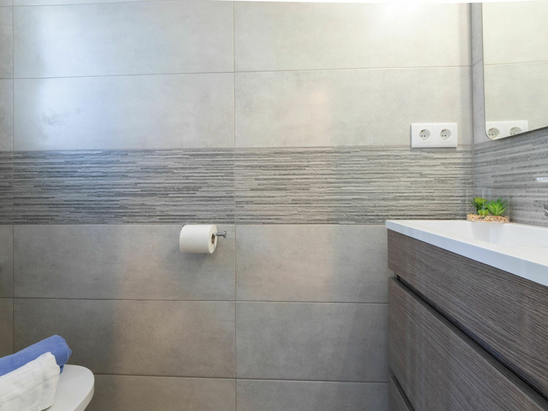 A bathroom with grey tiles, a toilet, a sink with a mirror, and a towel rack.