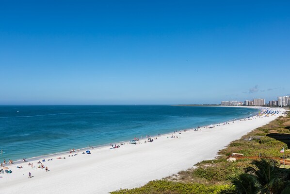 A wide beach with clear blue water, a white sandy shore, and a city skyline in the background.