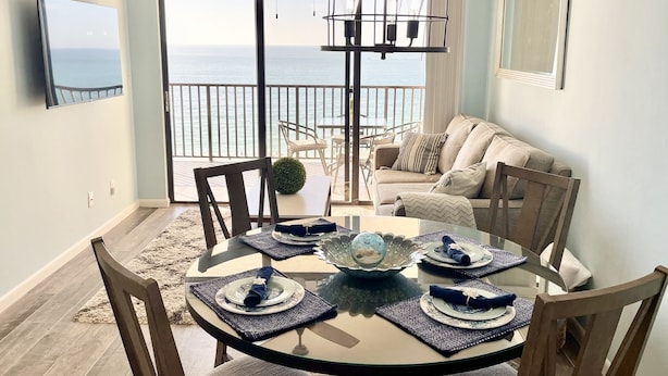 A dining area with a round table set for four, a balcony with a view of the sea, and a ceiling fan. - Upscale Sea Winds 8th Floor Beachfront Weekly Rental (Marco Island)