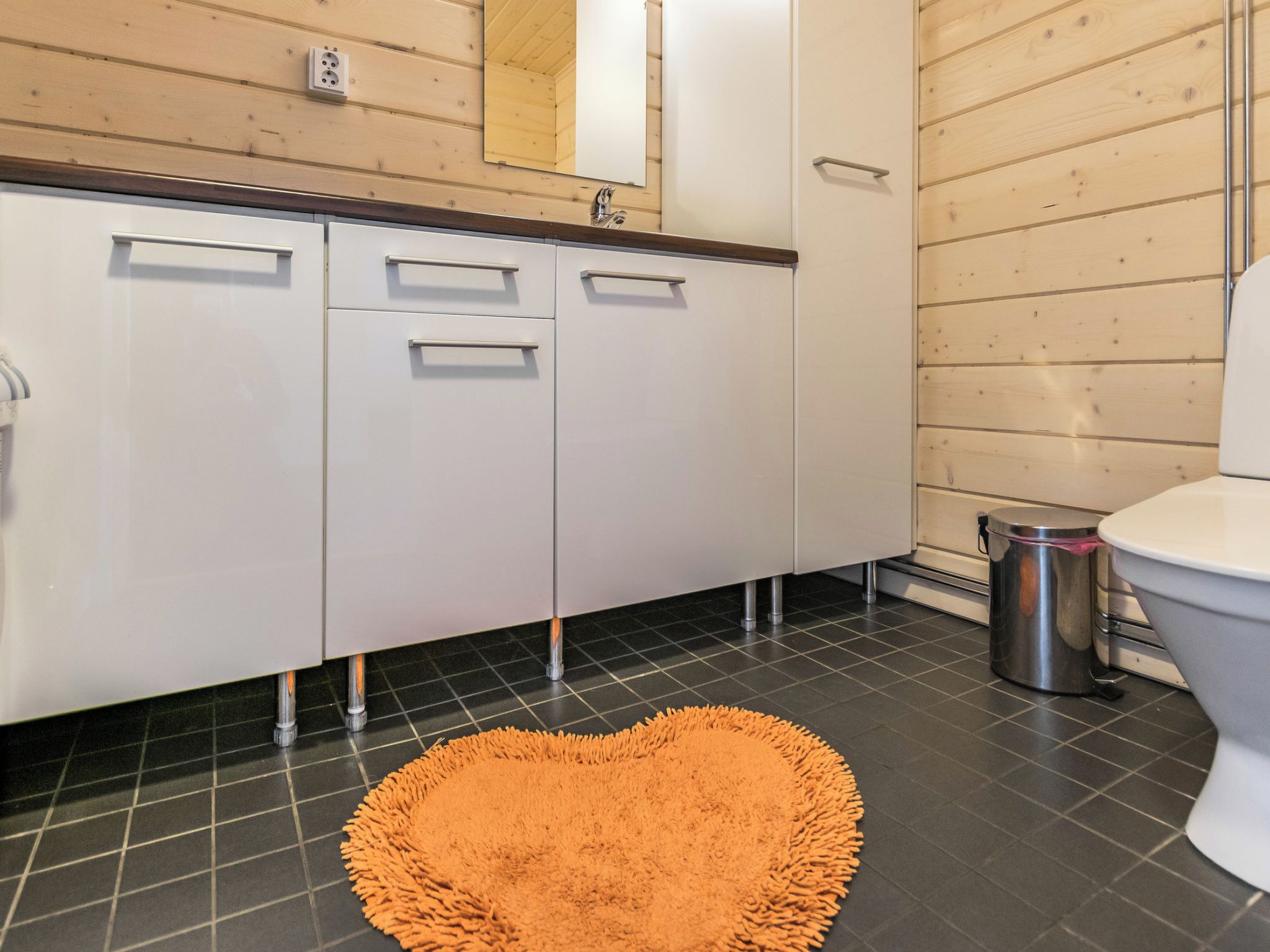 Une salle de bain moderne avec un lavabo blanc, un miroir et des toilettes. Le sol est recouvert de carreaux noirs et il y a un tapis orange.