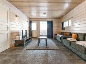 A room with a wooden ceiling, a television on a stand, a sofa with cushions, a table, and a window with curtains. - Hallan karhu c, paritalo by Interhome (Hyrynsalmi)