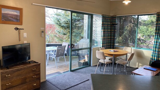 A room with a television, wooden dresser, and a dining table with chairs. There is a sliding glass door leading to an outdoor area with a patio and a view of trees. - Cosy cabin (Lumsden)