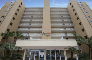A multi-story hotel with a central entrance, balconies on each floor, and palm trees in front. - Edgewater West 53 (Gulf Shores)