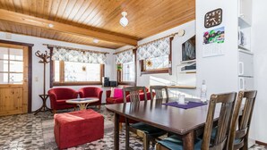 Ein gemütliches Zimmer mit Holzbalkendecke, einem Esstisch mit Stühlen, einem roten Sofa, einem roten Hocker und einem gemusterten Boden.