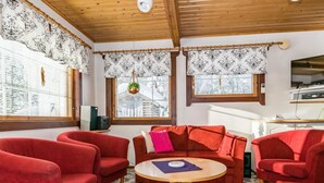 Ein gemütliches Wohnzimmer mit roten Sofas, einem Holztisch, gemusterten Vorhängen und Blick ins Freie.