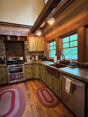A kitchen with a stone fireplace, wooden cabinets, a stainless steel stove, and a large window. - Creekside Fishing at TroutHaven – Paradise in the Mountains (McCaysville)