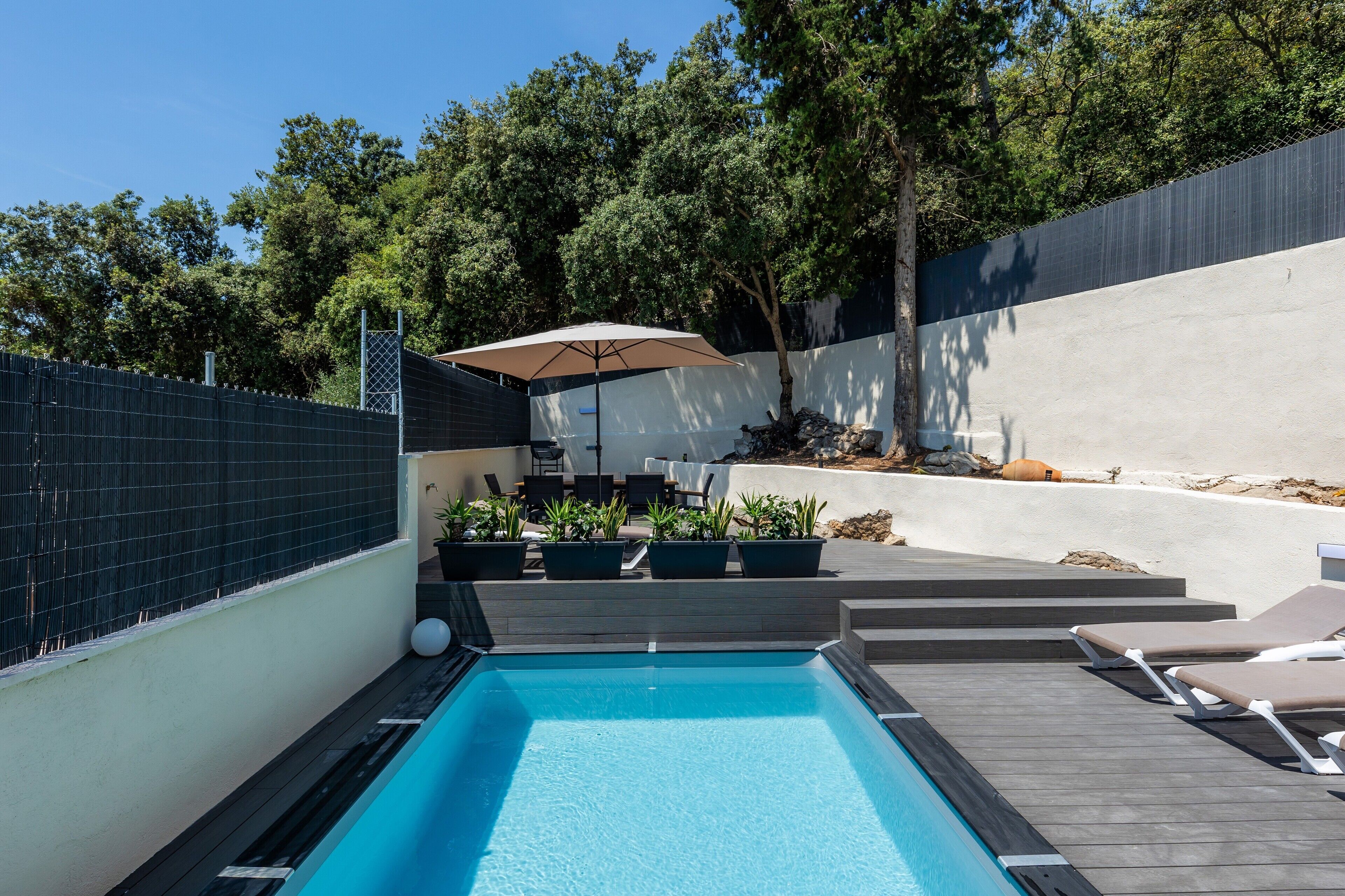 Una piscina con sillas de descanso, una mesa con sombrilla y plantas en macetas.