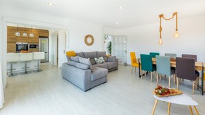 A modern living room with a grey sofa, a dining area with colorful chairs, and a kitchen with white appliances. - Vacation Home Sestel with Sea View, Pool, Balcony, Garden & Terrace (Capdepera)