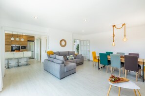 A modern living room with a grey sofa, a dining area with colorful chairs, and a kitchen with white appliances.