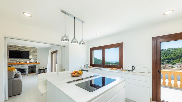 A modern kitchen with a central island, a built-in oven, and a large window with a view of the countryside. - Vacation Home Sestel with Sea View, Pool, Balcony, Garden & Terrace (Capdepera)