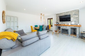 A modern living room with a grey sofa, a dining area with colorful chairs, a fireplace, and a flat-screen TV mounted on a stone wall.
