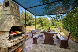 Une terrasse extérieure dotée d’un barbecue en pierre, de bancs en bois et d’une table dressée pour les repas.