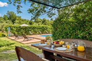 Un espace extérieur aménagé pour prendre les repas, avec une table dressée pour le petit-déjeuner, une piscine et des chaises longues en arrière-plan.