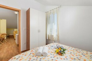 Une chambre à coucher avec un lit, une armoire, un rideau, une table avec un magazine et des fleurs, et une chaise.
