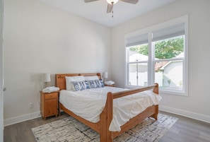 A bedroom with a wooden bed, white bedding, a nightstand with a lamp, and a large window with blinds.